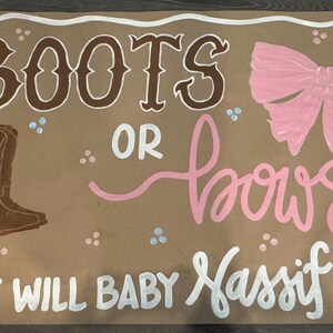 May include: A brown paper sign with the words "BOOTS OR BOWS?" in brown and pink lettering. The sign also includes a drawing of a cowboy boot and a pink bow. The text "WHAT WILL BABY Nassif BE?" is written in white.