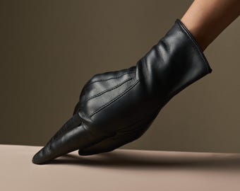Puede incluir: Primer plano de un guante de cuero negro. El guante es liso y se ajusta a la forma de la mano, con detalles de costura visibles en el dorso. Descansa sobre una superficie clara, con un fondo neutro.
