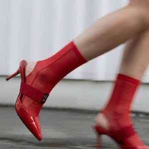 Puede incluir: Un par de zapatos de tac&oacute;n destalonados de charol rojo con punta afilada y tac&oacute;n fino. Los zapatos se llevan con calcetines rojos transparentes al tobillo que incluyen una banda el&aacute;stica roja con el texto TO BE impreso en color blanco.