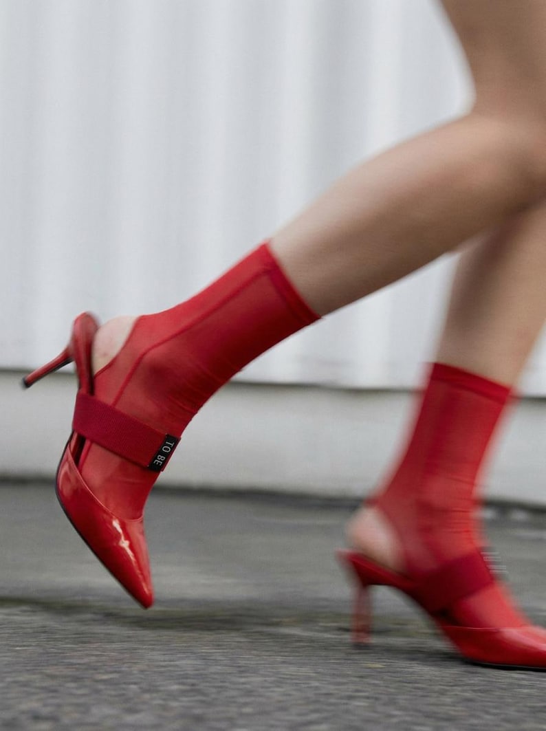 Puede incluir: Un par de zapatos de tac&oacute;n destalonados de charol rojo con punta afilada y tac&oacute;n fino. Los zapatos se llevan con calcetines rojos transparentes al tobillo que incluyen una banda el&aacute;stica roja con el texto TO BE impreso en color blanco.