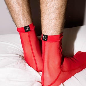 May include: Red sheer socks with the words "TO" and "BE" on the cuffs. The socks are worn on a person's feet, resting on a white bed sheet. The socks are a vibrant red color.