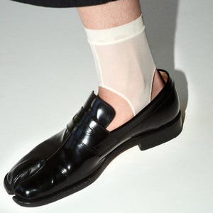 Puede incluir: Mocasines negros de cuero con puntera dividida y acabado brillante. Los zapatos tienen un tacón bajo y un diseño único, combinados con calcetines tobilleros blancos. Los mocasines son una versión moderna de un estilo clásico.