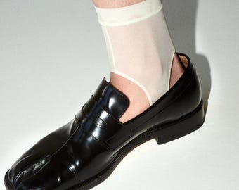 Puede incluir: Mocasines negros de cuero con puntera dividida y acabado brillante. Los zapatos tienen un tacón bajo y un diseño único, combinados con calcetines tobilleros blancos. Los mocasines son una versión moderna de un estilo clásico.