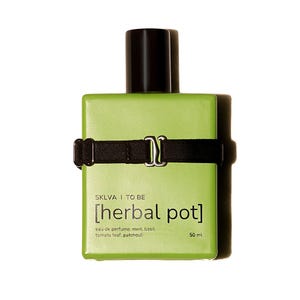 Op de afbeelding: Een vierkante, lichtgroene parfumfles met een zwarte dop en een zwarte band met een zilveren gesp. Het etiket luidt "SKLVA I TO BE [herbal pot] eau de parfum: munt, basilicum, tomatenblad, patchouli" en "50 ml".