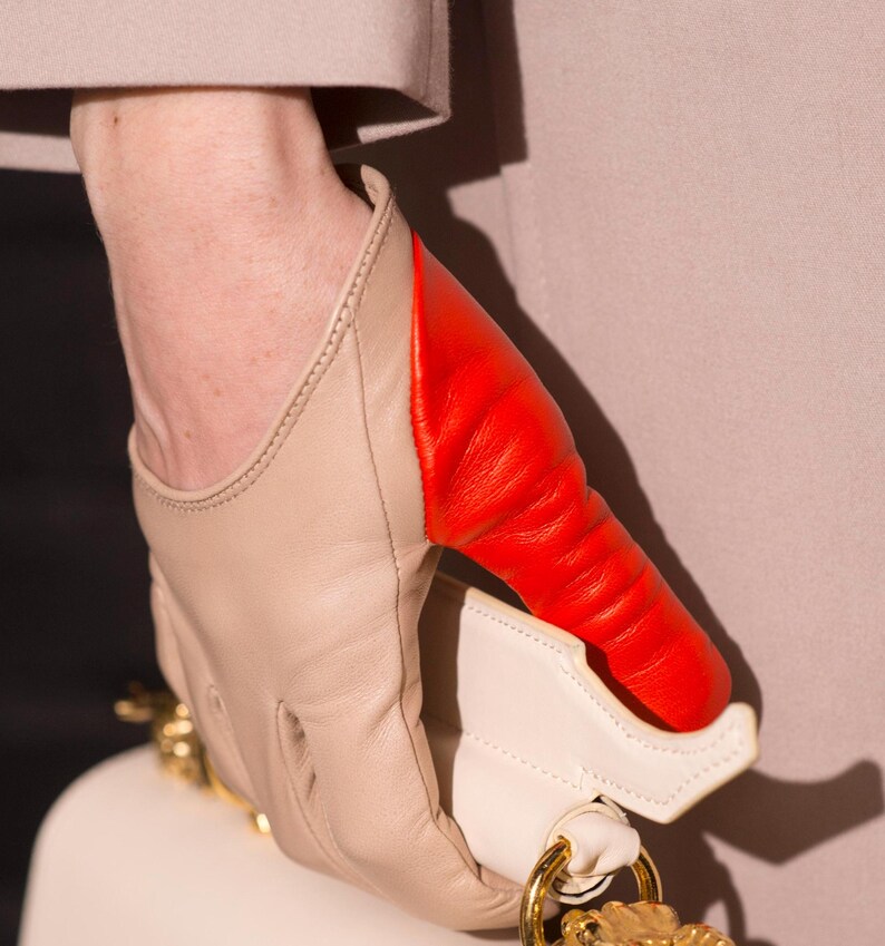 May include: A pair of leather gloves in a beige and red colour scheme. The gloves are made from soft, supple leather and have a unique design with a red panel on the back of the hand.