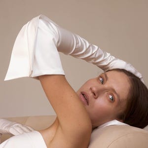 May include: A woman wearing a white satin long glove, lying on a cream-coloured couch. She is wearing a white strapless top and has her hair pulled back.