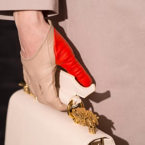 May include: A white leather handbag with a gold lion head clasp. The handbag is being held by a hand wearing a beige and red leather glove.