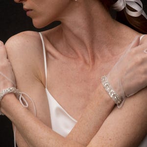 K&ouml;nnte beinhalten: Eine Frau tr&auml;gt ein wei&szlig;es Satin-Slip-Kleid mit einem wei&szlig;en Band im Haar. Sie tr&auml;gt an jeder Hand ein Paar mit Kristallen verzierter Handschuhe.