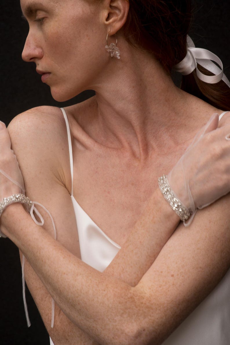 K&ouml;nnte beinhalten: Eine Frau tr&auml;gt ein wei&szlig;es Satin-Slip-Kleid mit einem wei&szlig;en Band im Haar. Sie tr&auml;gt an jeder Hand ein Paar mit Kristallen verzierter Handschuhe.