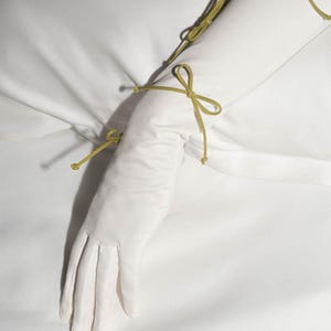 Op de afbeelding: Lange witte handschoenen met groene lintjes. De handschoenen zijn met strikken aan de pols en elleboog gebonden.