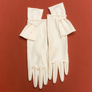 Puede incluir: Un par de guantes de color marfil con un diseño clásico, con un detalle de puño con volantes. Los guantes están hechos de una tela lisa y están colocados sobre un fondo liso de color marrón rojizo.