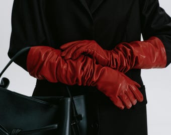 Op de afbeelding: Een persoon draagt een zwarte jas en lange rode leren handschoenen en houdt een zwarte leren handtas vast met een geknoopte handgreep.