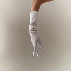Puede incluir: Un guante largo de satén blanco. El guante se extiende más allá de la muñeca y está ligeramente fruncido en la parte superior. Los dedos son delgados y la tela parece suave. El fondo es de color blanco roto neutro.