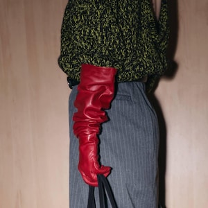 May include: A long, red leather glove with a ruffled design. The glove is held by a person wearing a black and green sweater and gray pinstripe pants. The glove is the focal point of the image.