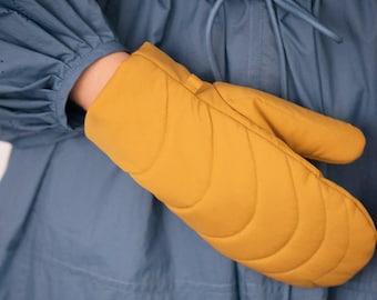 Puede incluir: Manopla de horno acolchada de color amarillo mostaza. El guante tiene un patrón acolchado curvo y se muestra sobre un fondo azul. El guante está diseñado para proteger las manos del calor al manipular utensilios de cocina calientes.