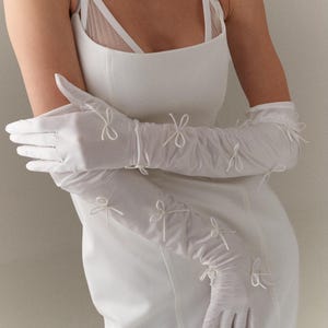 Op de afbeelding: Witte, ellebooglange handschoenen met delicate strikaccenten. De handschoenen worden gecombineerd met een witte jurk met een vierkante halslijn en spaghettibandjes. De strikken zijn gelijkmatig verdeeld.