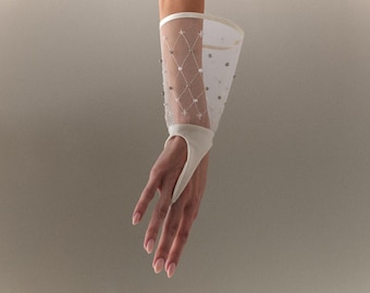 Puede incluir: Guante de novia blanco con un lazo para el dedo y un puño transparente y acampanado. El puño está adornado con un patrón de diamantes de pequeñas perlas y detalles plateados. El guante está hecho de una tela suave y delicada.