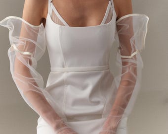 May include: A white bridal gown with a square neckline and a fitted bodice. The dress features sheer, long sleeves with a ruffled edge and matching sheer gloves. A thin belt cinches the waist. The overall style is elegant and minimalist.