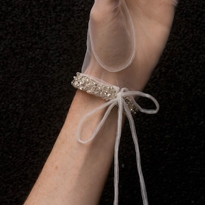 K&ouml;nnte beinhalten: Wei&szlig;e T&uuml;llhandschuhe mit einem perlenbesetzten Handgelenksmanschetten und einer Schleife. Die Handschuhe sind durchsichtig und zart.