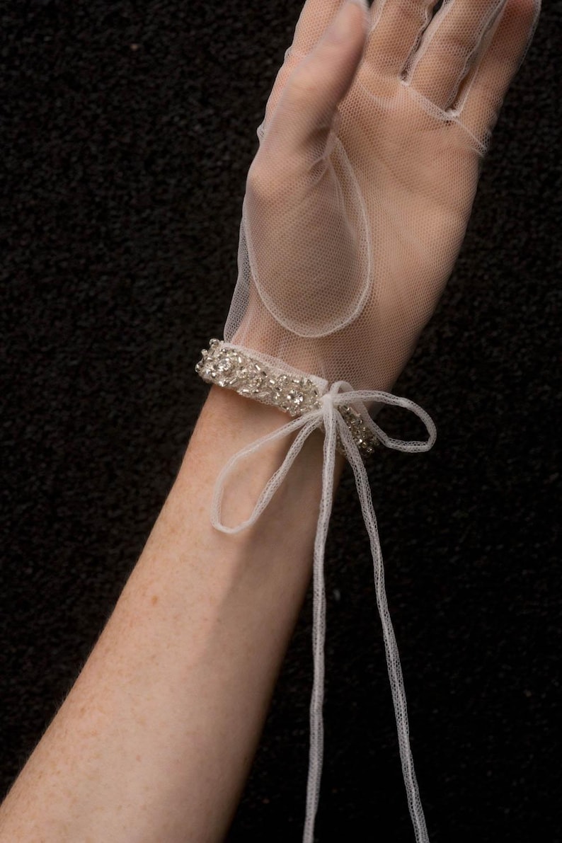 K&ouml;nnte beinhalten: Wei&szlig;e T&uuml;llhandschuhe mit einem perlenbesetzten Handgelenksmanschetten und einer Schleife. Die Handschuhe sind durchsichtig und zart.
