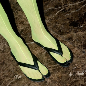 May include: A pair of black split-toe thong sandals worn with sheer neon yellow socks. The sandals feature a distinct separation between the big toe and the other toes, with a smooth matte finish on the straps and footbed, paired with bright hosiery.
