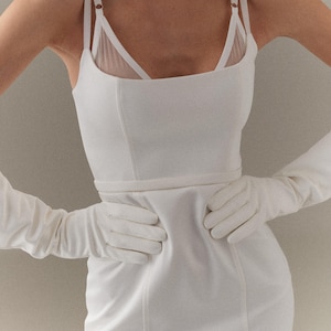 Könnte beinhalten: Ein weißes Kleid mit quadratischem Ausschnitt und dünnen Trägern. Das Kleid hat ein figurbetontes Oberteil und einen geraden Rock. Lange weiße Handschuhe werden getragen, die Hände auf der Taille. Ein durchsichtiger weißer BH ist sichtbar.