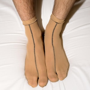 May include: Tan socks with a split toe design, featuring a black stitched seam down the front. The socks have a cuff at the top and are worn on a white surface.