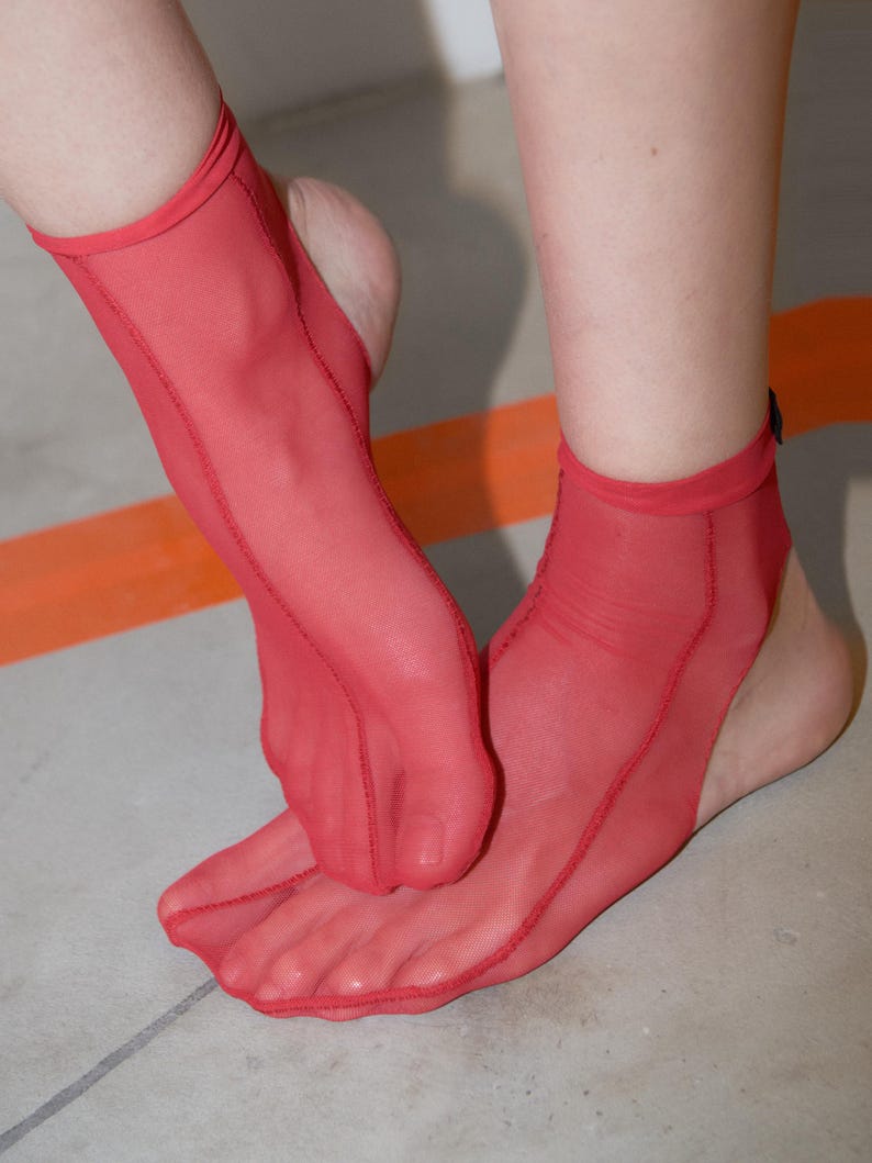 Puede incluir: Un par de calcetines tobilleros rojos transparentes con dise&ntilde;o de dedos separados. Los calcetines est&aacute;n confeccionados con un tejido de malla fino y transl&uacute;cido con costuras visibles a lo largo de la parte superior y los lados, dejando el tal&oacute;n al descubierto.