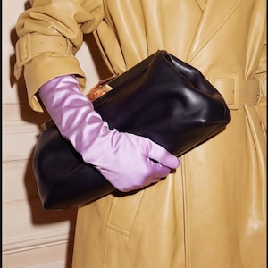 May include: A black leather clutch is held by a person wearing long, light purple satin gloves. The person is wearing a tan leather trench coat with a matching belt and buckle. The image is a fashion shot.