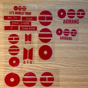 May include: A collection of red vinyl decals featuring the BTS logo, the text BTS WORLD TOUR, the names RM, JIN, SUGA, JIMIN, J-HOPE, V, and JUNG KOOK, and the word ARIRANG. The set includes various circular designs with horizontal cutouts.