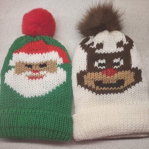 May include: Two knitted winter hats. One is green with a Santa Claus face and a red pom-pom. The other is white with a reindeer face and a brown pom-pom. Both hats have a folded brim.