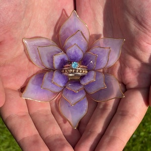 Op de afbeelding: Een paarse en gouden bloemenringhouder met een blauwe edelsteenring erin. De houder is gemaakt van hars en draad.