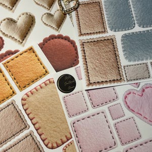 May include: A collection of felt stickers in various shapes and colors, including hearts, squares, and rectangles. The stickers feature stitched edges and a soft, textured appearance. Two gold heart-shaped charms are also visible.
