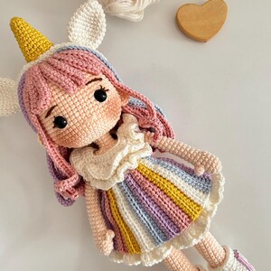 Puede incluir: Una muñeca de ganchillo hecha a mano con forma de unicornio. La muñeca tiene un cuerno amarillo, orejas blancas y pelo rosa, morado y azul. La muñeca lleva un vestido a rayas arcoíris y botas blancas. Un corazón de madera y lana en el fondo.