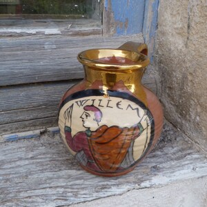 Puede incluir: Una jarra de cerámica marrón con un borde dorado y una imagen pintada de una persona con la palabra "WILLEM" en el lateral.