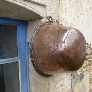 May include: A large copper pot hangs on a white wall with a blue painted window. The pot has a shiny, metallic finish and is slightly dented.