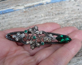 May include: A silver-toned brooch with a floral design featuring green and clear rhinestones. The brooch is shaped like a long oval and has a large green rhinestone at one end.