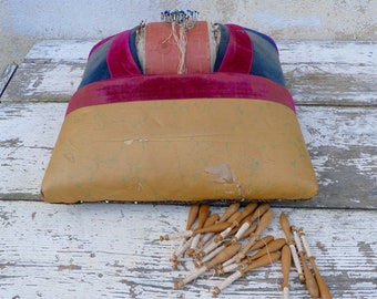 May include: A vintage pincushion with a brown, blue, and pink velvet cover. The pincushion is filled with pins and is sitting on a white wooden surface. There are also a number of wooden bobbins with white thread on them.
