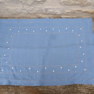 May include: A rectangular, pale blue placemat with a white dotted border and a stitched rectangular design. The placemat is made of a soft fabric and is on a wooden surface.
