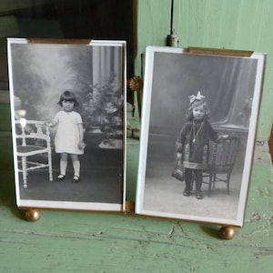 Puede incluir: Marco de fotos de doble cara con dos retratos en blanco y negro de niños. El marco es de latón y cristal, con un acabado dorado. Descansa sobre dos pequeños pies redondos.