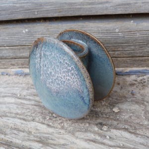 Peut inclure: Un bougeoir en céramique fait à la main. Il présente deux assiettes ovales émaillées en bleu avec une surface texturée et un bord marron. Les assiettes sont reliées par une petite poignée incurvée. Objet décoratif pour bougies.