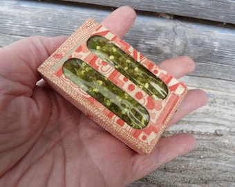 May include: A small, rectangular box with a decorative red and cream pattern. The box contains two clear tubes filled with green and yellow beads. The box is held in a hand.