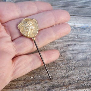 Puede incluir: Alfiler de sombrero dorado con un perfil femenino clásico. El alfiler tiene una cabeza decorativa con un relieve detallado y un largo vástago de metal oscuro. Probablemente vintage o antiguo.