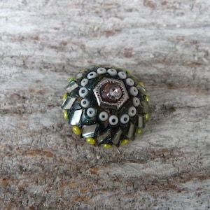 Könnte beinhalten: Ein handgefertigter Knopf im Mosaikstil mit gewölbter Form. Der Knopf hat ein zentrales, silberfarbenes, sechseckiges Element mit einem rosa Edelstein. Das Design umfasst dunkelgrüne, silberne, weiße und gelbe Akzente, die einen strukturierten, dekorativen Effekt erzeugen.