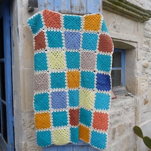Peut inclure: Une couverture au crochet faite à la main avec un motif patchwork. La couverture présente des carrés aux tons de turquoise, orange, jaune, bleu et marron, avec une bordure crème. La couverture est drapée sur un volet en bois bleu vieilli.