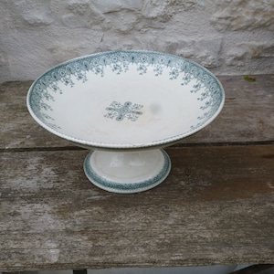Peut inclure: Un support à gâteau en céramique blanc avec un motif floral vert. Le support a une base ronde et un bol large et peu profond.
