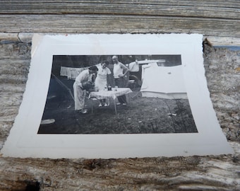 May include: Black and white photo of four people gathered around a table outdoors. They are sitting and standing, and appear to be enjoying a meal together. There is a car and a tent in the background.