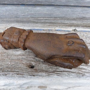 Può includere: Una mano in ghisa arrugginita con una lettera "D" in rilievo sul retro della mano. La mano è chiusa a pugno.