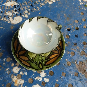 May include: A white teacup with a green and brown floral design on the saucer. The teacup and saucer are on a blue metal table.