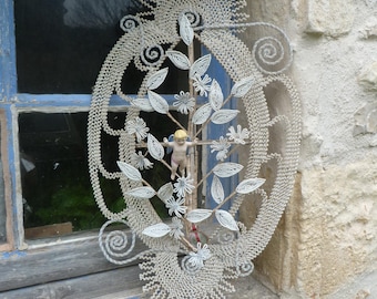 May include: An ornate, handmade wall hanging with a central design of a small cherub figure and a beaded floral motif. The piece is primarily white with intricate detailing, set against a weathered wooden window frame and stone wall.